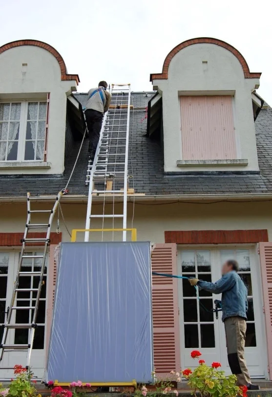 Monte-meuble à échelle posé au sol contre la façade d'une maison, montant un grand panneau emballé jusqu'à la toiture